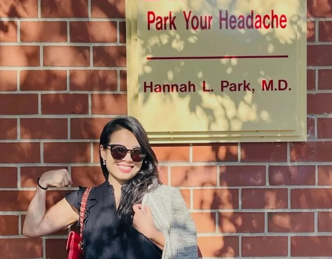 Dr. Hannah Park smiling and flexing her arm beneath the Park Your Headache office sign in Voorhees
